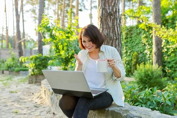 Beautiful emotional middle aged woman relaxing in garden with cup of tea and laptop