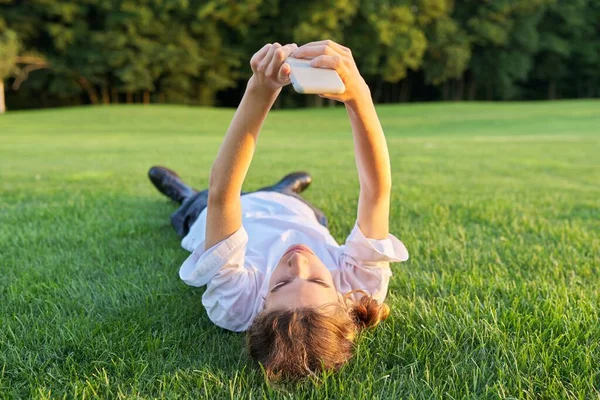 10代の若者がスマートフォンを持って草の上に横たわっている。男性で白いTシャツに電話で彼の手、トップビュー、緑の芝生の背景 — ストック写真