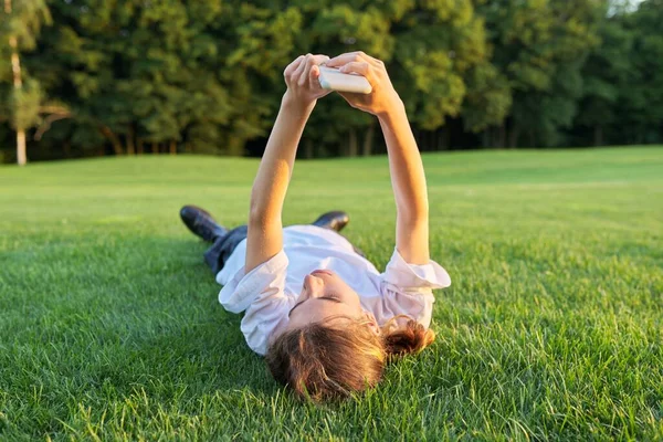 En tonåring som ligger på gräs med smartphone. Man i vit T-shirt med telefon i händerna, ovanifrån, grön gräsmatta bakgrund — Stockfoto