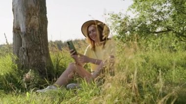 Gülümseyen, konuşan bir kadın, çayırda oturmuş akıllı telefon ekranına bakıyor. Video çağrısı kullanarak.