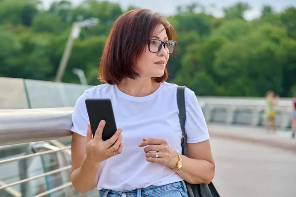 Portrait of middle-aged woman walking with smartphone in hand, summer day in city — Stock Photo, Image