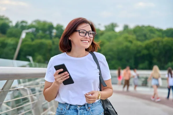 Portrait of middle-aged woman walking with smartphone in hand, summer day in city — Stock Photo, Image