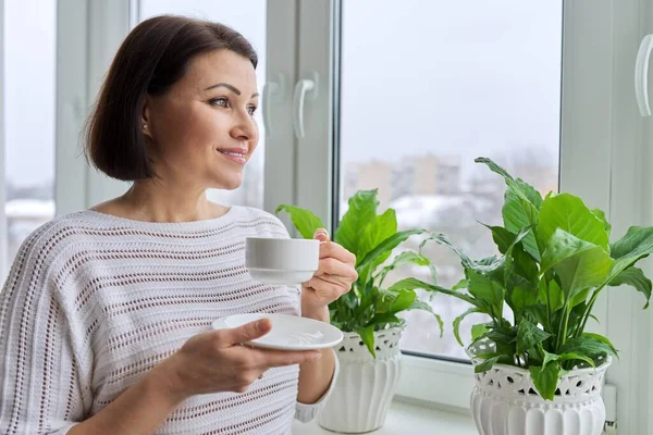 Temporada invierno, día nevado, mujer sonriente de mediana edad con taza de café mirando por la ventana. — Foto de Stock