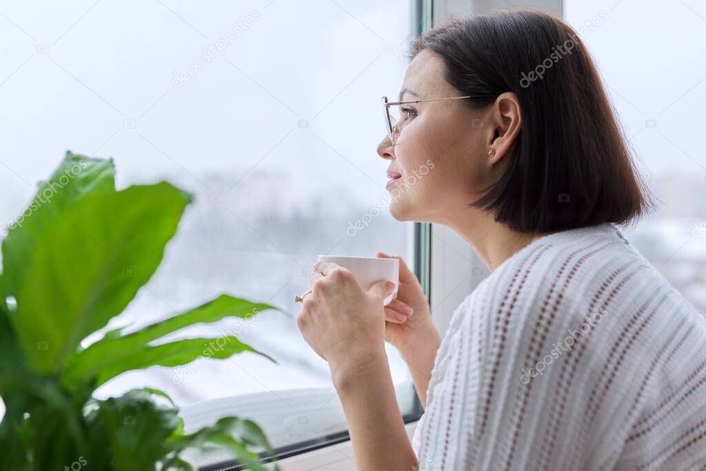 Season winter, snowy day, middle aged smiling woman with cup of coffee looking out the window.