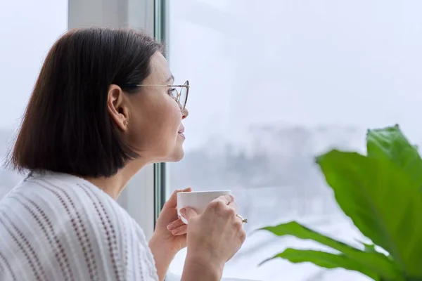 Temporada de invierno, nieve, mujer de mediana edad mirando a la ventana del hogar — Foto de Stock