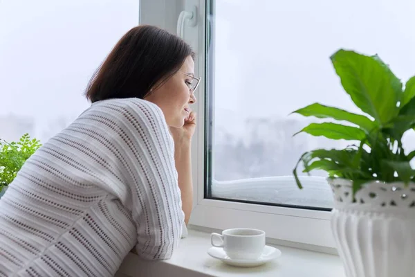 Зимний сезон, снег, женщина средних лет смотрит в окно дома — стоковое фото