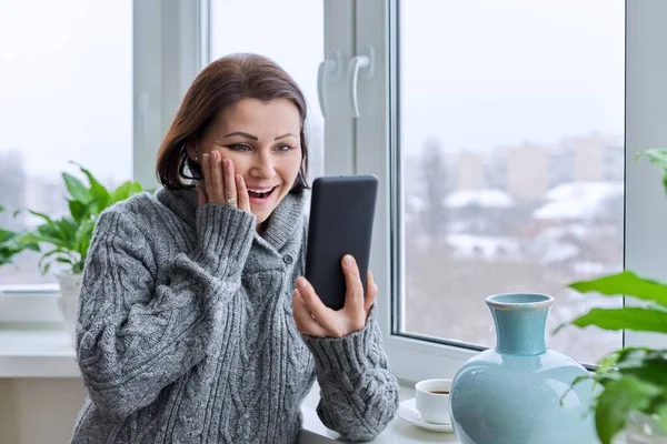 Surprised happy middle aged woman in warm knitted sweater with smartphone in hands — ストック写真