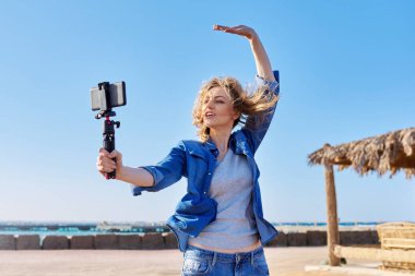 Akıllı telefonlu orta yaşlı bir kadın web kamerasına bakıyor ve video kaydediyor.