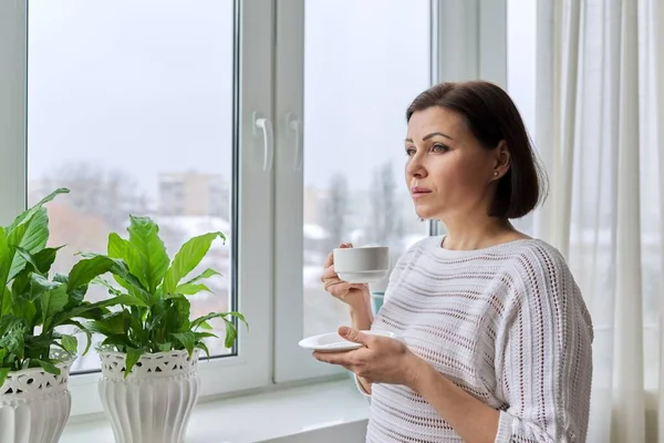 Allvarlig medelålders kvinna tittar ut genom fönstret — Stockfoto