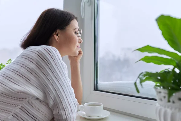 Temporada de invierno, nieve, mujer de mediana edad mirando a la ventana del hogar —  Fotos de Stock