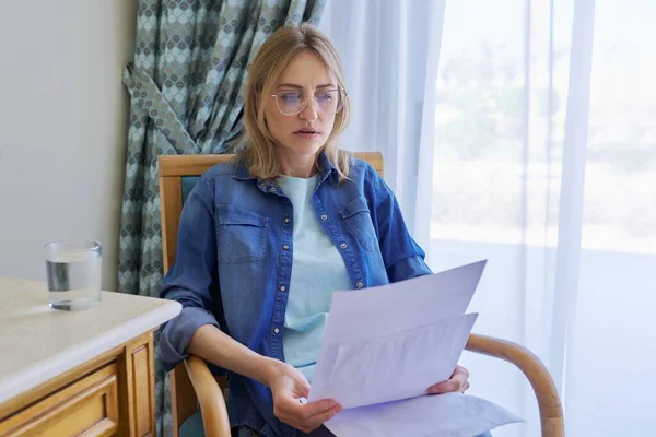 Mujer seria de mediana edad con gafas leyendo papeles sentados en casa en silla —  Fotos de Stock