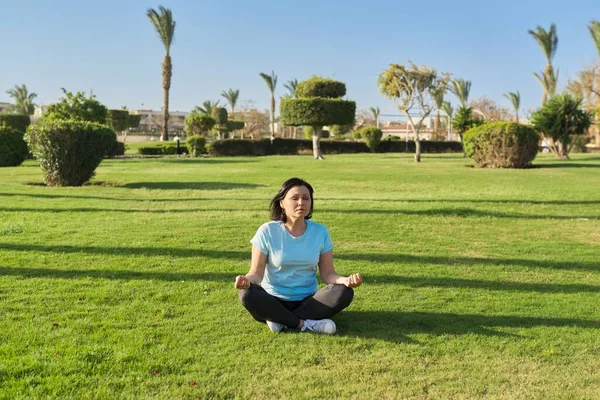 Middle aged woman sitting on grass in lotus position and meditating with closed eyes — Stock Photo, Image