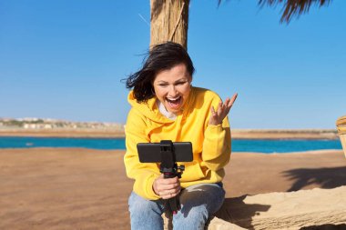 Orta yaşlı bir kadın akıllı telefondan, deniz tatilinden, doğadan, deniz manzarasından video kaydediyor.