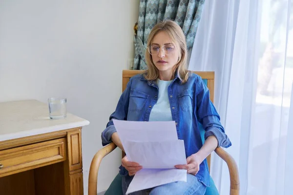 Seriöse Frau mittleren Alters mit Brille, die zu Hause im Stuhl Zeitung liest — Stockfoto