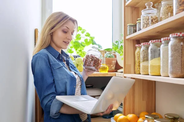Middle-aged woman in kitchen in pantry with laptop showing cans of food. Female food blogger, online communication, healthy eating video recording, healthy food, technology, hobbies and leisure