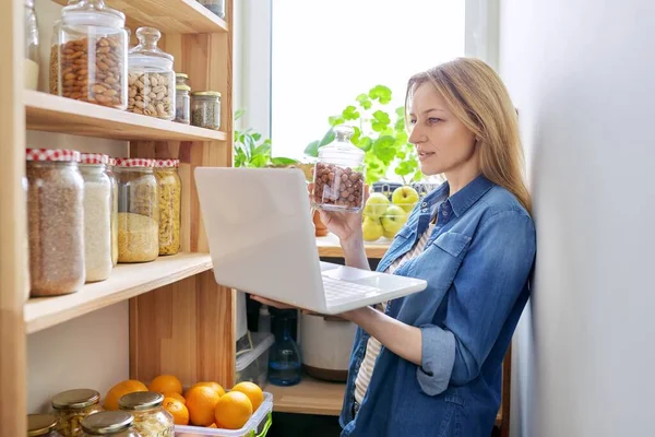 Mujer de mediana edad en cocina en despensa con laptop mostrando latas de comida — Foto de Stock