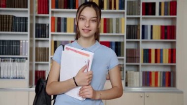 Kütüphanenin arka planında, kameraya bakan gülümseyen genç kız öğrenci portresi.