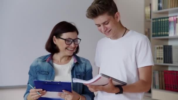В колледже 17-летний мальчик-подросток разговаривает с учительницей-наставницей в библиотеке класса — стоковое видео