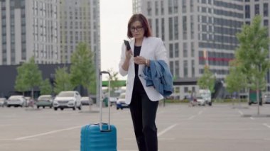 Bavullu orta yaşlı iş kadını açık havada cep telefonuyla konuşuyor.