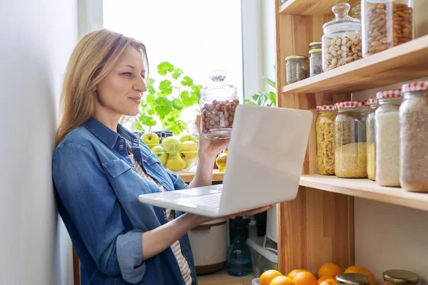 Mulher de meia-idade na cozinha em despensa com laptop mostrando latas de comida — Fotografia de Stock