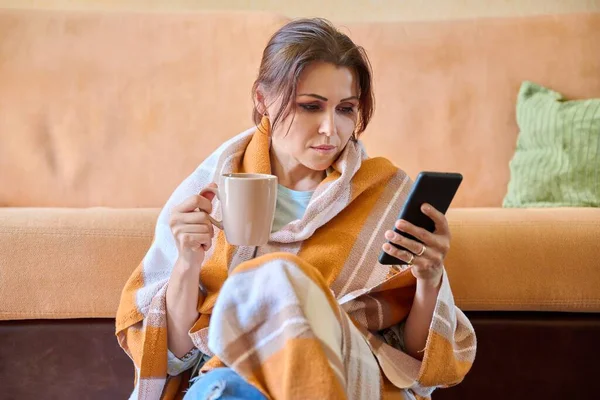 Herbst-Winter-Porträt einer Frau mittleren Alters unter warmer Decke mit Tasse und Smartphone — Stockfoto