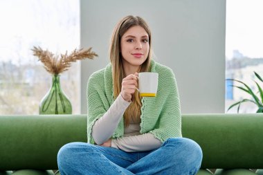 Evdeki kanepede elinde fincanla oturan rahat genç bir kadının portresi. Gülümseyen çekici bir kadın oturma odasında kupayla kanepede dinleniyor. Boş zaman mutluluğu rahat yaşam tarzı gençlik konsepti