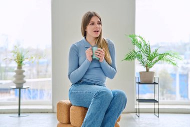 Rahatlamış genç bir kadının portresi, evinde elinde bardakla sandalyede oturuyor. Gülümseyen çekici bir kadın oturma odasında kupayla koltukta dinleniyor. Boş zaman mutluluğu rahat yaşam tarzı gençlik konsepti