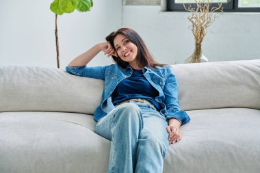 Oturma odasındaki kanepede oturan güzel, mutlu, esmer kadının portresi. 30 'lu yaşlarda çekici bir kadın kameraya bakıyor. Güzellik yaşam tarzı rahatla rahat rahat mutluluk, kadın konsepti