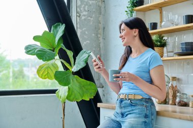 Güzel, mutlu, gülümseyen, 30 'lu yaşlardaki bir kadının portresi. Mutfakta akıllı telefon kullanan, profil manzaralı bir kadın. Yaşam tarzı, ev hayatı, insan kavramı.