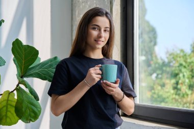 Gülümseyen, gevşemiş genç kızın portresi. Evde, pencerenin yanında poz veriyor. Kameraya bakan uzun saçlı güzel bir genç bayan. Güzellik, sağlık, yaşam tarzı, ev, gençlik kavramı