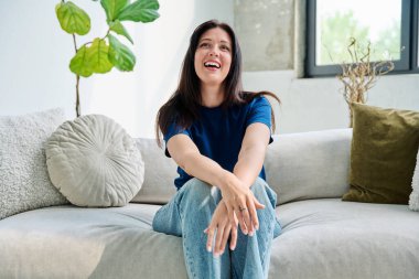 Oturma odasındaki kanepede oturan güzel, mutlu esmer kadının portresi. 30 'lu yaşlarda çekici bir kadın kameraya bakıyor. Güzellik, yaşam tarzı, kadın konsepti