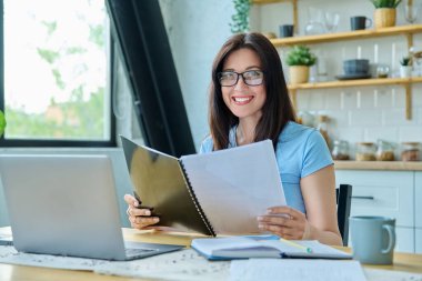 Dizüstü bilgisayarla çalışan gülümseyen genç bir kadın, iş gazetelerini okuyan, kameraya bakan, mutfaktaki masada oturan pozitif bir kadın. Teknoloji, iş, iş, 30 'ların insan kavramı