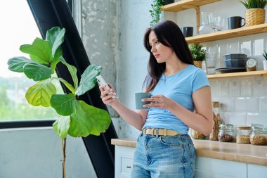 Güzel, mutlu, gülümseyen, 30 'lu yaşlardaki bir kadının portresi. Mutfakta akıllı telefon kullanan, profil manzaralı bir kadın. Yaşam tarzı, ev hayatı, insan kavramı.