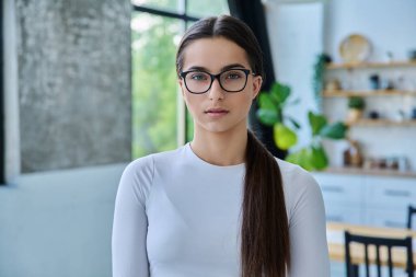 Gözlüklü, evde poz veren ciddi bir genç kızın portresi. Kameraya bakan uzun saçlı güzel bir genç bayan. Güzellik, sağlık, yaşam tarzı, gençlik kavramı