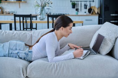 Evde dizüstü bilgisayarla kanepede uzanmış sınav için notlar alan genç üniversiteli kız. Eğitim e-öğrenim kursu online ders yaşam tarzı gençlik kavramı