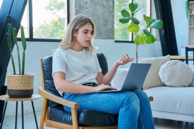 Genç ve güzel bir kadın internette sohbet konferansı için dizüstü bilgisayar kullanıyor. Evde, sandalyesinde oturuyor. İletişim öğrenmek için dijital teknolojiler sosyal medya konsepti bloglarında çalışıyor