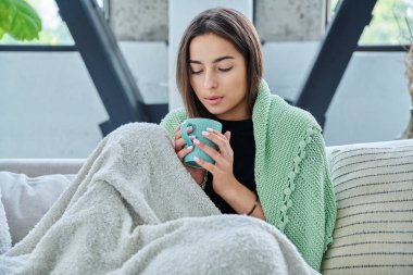 Oturma odasındaki kanepede elinde fincanla sıcak battaniyenin altında oturan genç ve güzel bir kadın. Kupaya bakan bir kız. Soğuk bahar sonbahar kış mevsimi, tatiller rahat yaşam tarzı konsepti