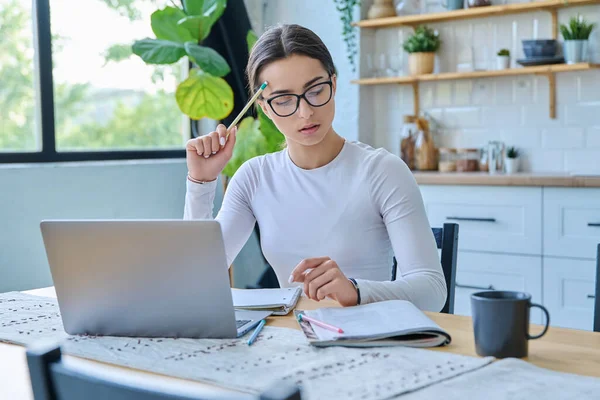 Masada dizüstü bilgisayarı, not defterleri ve ev içinde ders kitapları olan genç bir kız öğrenci oturuyor. Eğitim, teknoloji, gençlik kavramı