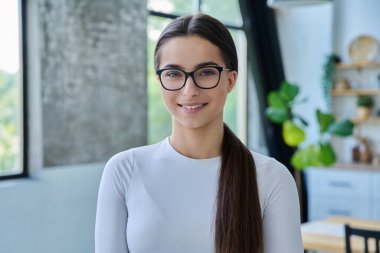 Gözlüklü, evde poz veren başarılı bir genç kızın portresi. Kameraya bakan uzun saçlı, gülümseyen genç bir bayan. Güzellik, sağlık, yaşam tarzı, gençlik kavramı