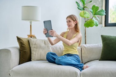 Genç ve güzel bir kız öğrenci dijital tabletle online sohbet konferansı yapıyor. Evde, kanepede oturuyor. İletişim öğrenme teknolojileri gençlerin video bloglarında boş zamanlarında çalışıyor