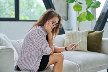 Üzgün, ciddi, orta yaşlı ve akıllı telefonlu bir kadın kanepesinde oturuyor. Kötü haberler, iş hayatında sorunlar, vücut ağrısı, akıl sağlığı konsepti