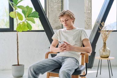 Evde oturan rahat genç adam akıllı telefon kullanarak sandalyede oturuyor. Dinlenme, rahatlama, rahatlama, ev hayatı, yaşam tarzı, ergenlik kavramı.