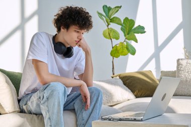 Genç yakışıklı kıvırcık adam internette sohbet etmek için bilgisayarını kullanıyor. Evde, koltukta oturuyor. İletişim öğrenimi için dijital teknolojiler boş zamanlarında sosyal medya bloglarında çalışır