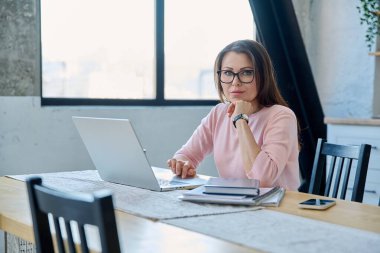 Orta yaşlı gülümseyen güzel bir kadın dizüstü bilgisayar belgeleriyle çalışıyor, mutfaktaki masada oturan kameraya bakıyor. Teknoloji, iş, iş, olgun insanlar konsepti