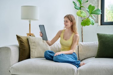 Genç ve güzel bir kız öğrenci dijital tabletle online sohbet konferansı yapıyor. Evde, kanepede oturuyor. İletişim öğrenme eğitimi için dijital teknolojiler boş zamanlarında blog yazarlığı