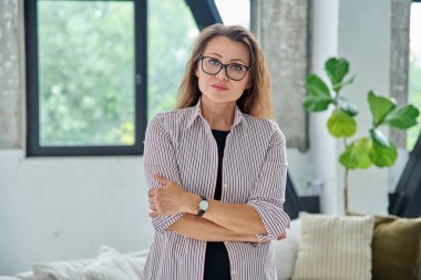 Kollarını kavuşturup poz veren kendinden emin orta yaşlı bir kadın portresi. Ofiste başarılı ve olgun bir bayan.