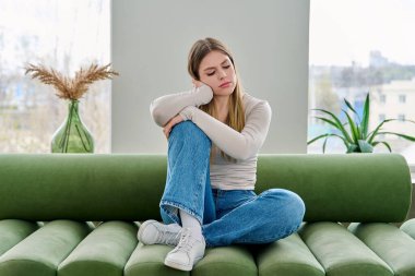 Evdeki kanepede oturan mutsuz, üzgün genç bir kadın, yüzü yakın çekim. Duygusal fiziksel acı çeken üzgün bir kadın. Sorunlar ruhun ve bedenin sağlığı, gençlik kavramı