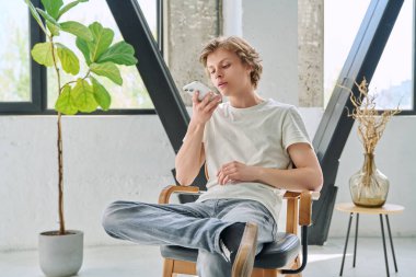 Evde oturan rahat genç adam sesli mesaj göndermek için akıllı telefonu kullanıyor. Dinlenme, rahatlama, rahatlama, ev hayatı, yaşam tarzı, ergenlik kavramı.