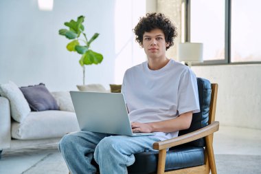 Evdeki sandalyede oturan dizüstü bilgisayarı olan yakışıklı, ciddi bir erkek portresi. 20 yaşında, kameraya bakan kıvırcık bir öğrenci. Yaşam tarzı, eğitim, teknoloji, gençlik kavramı