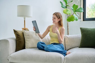 Genç ve güzel bir kız öğrenci dijital tabletle online sohbet konferansı yapıyor. Evde, kanepede oturuyor. İletişim öğrenme teknolojileri gençlerin video bloglarında boş zamanlarında çalışıyor